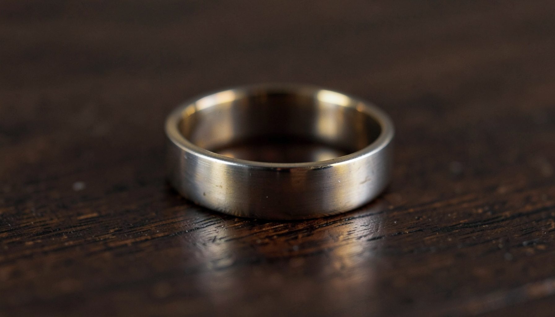 A softly lit wedding band resting on a dark wooden surface, with muted gold and silver tones against deep brown, evoking reflection and quiet emotion