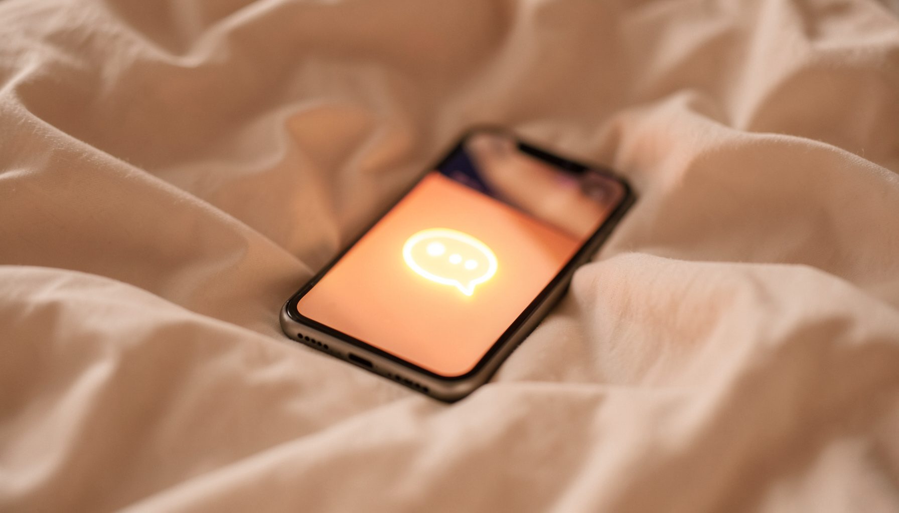 A softly lit smartphone resting on rumpled cream bedsheets, its screen glowing with a message bubble icon, warm amber tones creating an intimate evening feel