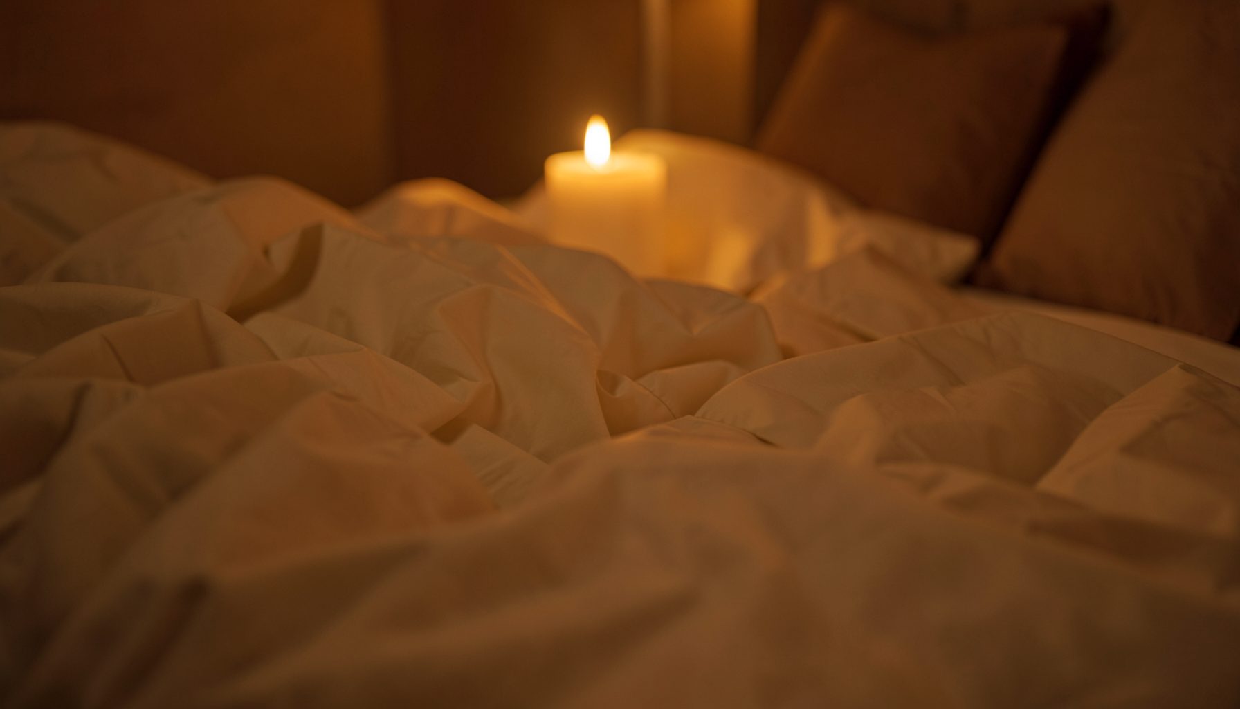 Soft candlelit bedroom scene with rumpled cream sheets and warm amber lighting, creating an intimate and relaxed atmosphere